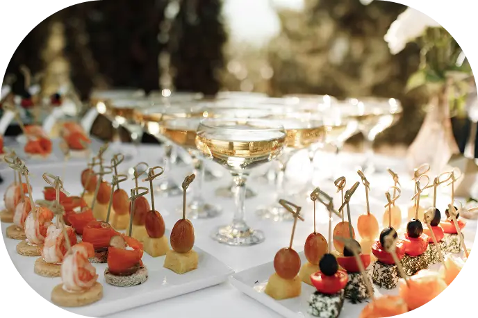 Elegancka aranżacja ze szklankami do szampana i różnorodnymi przekąskami na szaszłykach, w tym krewetkami, pomidorami i oliwkami, na oświetlonym słońcem stole.