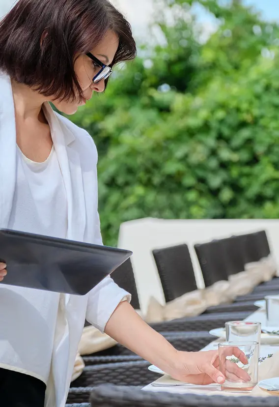 Kobieta w białej marynarce układa szklanki na długim, eleganckim stole ogrodowym, nakrytym świecami i talerzami, sprawiając wrażenie osoby przygotowanej i uważnej.